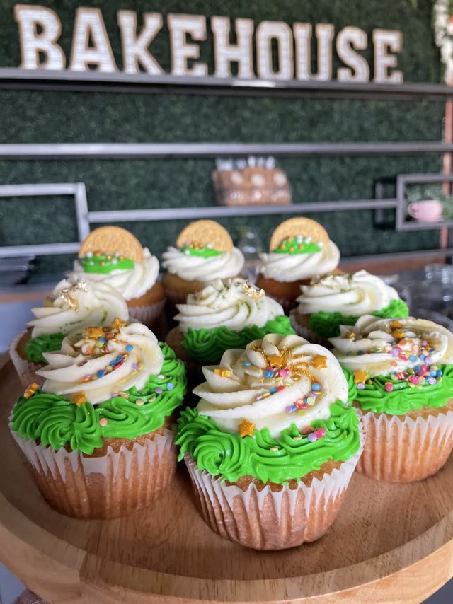 Cupcakes with green and cream swirled frosting, rainbow sprinkles and gold stars on a wooden stand, in front of a green wall with the Bakehouse sign