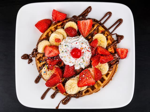 A waffle on a white plate topped with strawberries, banana slices, chocolate drizzle, whipped cream with sprinkles and a cherry