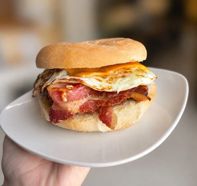 A hand holding a plate with a bagel sandwich of bacon, fried egg and melted cheese
