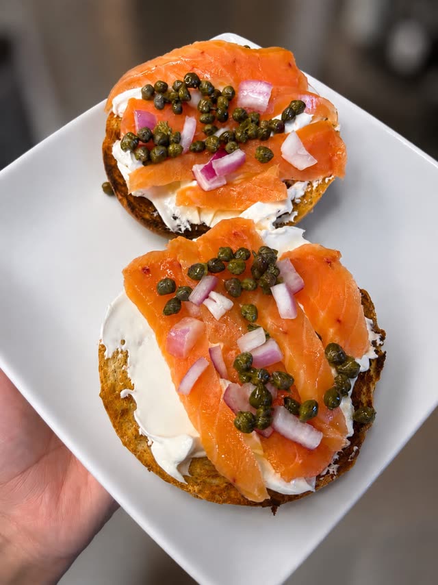 A hand holding a plate of two bagel halves with cream cheese, lox, capers and red onion