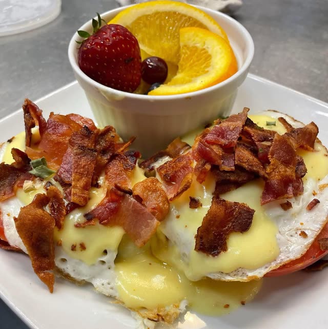 Fried eggs topped with hollandaise and chopped bacon, served beside a cup of fruit with a strawberry and orange slices