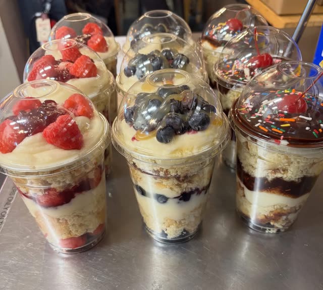 Nine layered cake cups with clear lids, topped with raspberries, blueberries, or chocolate with sprinkles and a cherry